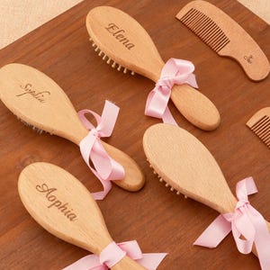 Puede incluir: Cepillos y peines de madera para bebés, con nombres grabados y lazos rosas. Los cepillos tienen cerdas suaves. Los peines también son de madera y tienen un diseño sencillo.