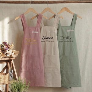 May include: Three personalized aprons in pink, beige, and green hang on a wooden rod. The aprons have pockets and are labeled with names and phrases: "Yoliana", "Jessica Made With Love", and "Emily Born to Garden". A small wooden ladder holds gardening tools and dried flowers.