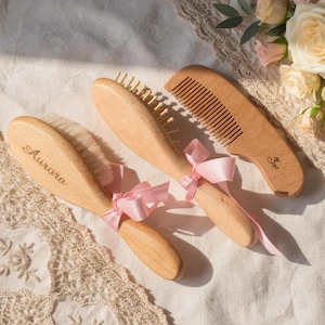 Puede incluir: Tres artículos de cuidado del cabello de madera: dos cepillos y un peine. Los cepillos tienen cerdas de color claro y están atados con cintas rosas. Un cepillo está grabado con el nombre "Aurora". El peine también es de madera y tiene un mango.