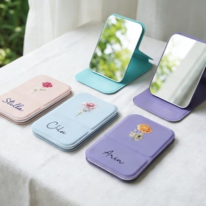 Puede incluir: Una colección de espejos compactos en varios colores pastel, incluyendo rosa, azul, turquesa y morado. Cada espejo presenta un diseño floral y un nombre, como "Stella", "Chloe" y "Aria". Los espejos se muestran abiertos y cerrados.