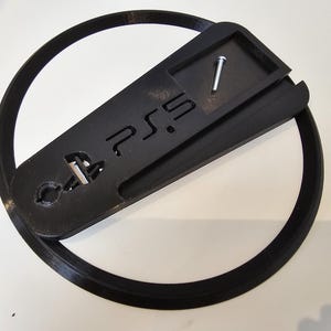 May include: A black PS5 console stand with a circular base. The stand has the PS5 logo and a slot for the console. A silver screw is included. The stand is designed to hold the console securely and provide stability.