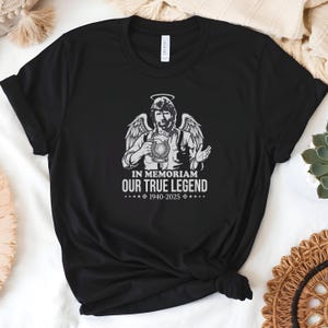 May include: Black t-shirt featuring a graphic of a man with angel wings, a halo, and a beer stein. The text "IN MEMORIAM OUR TRUE LEGEND 1940-2025" is printed below the image. The shirt is made of a soft material.