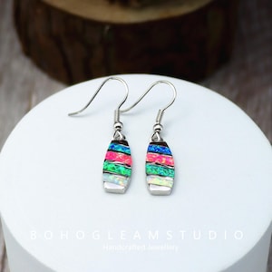 May include: A pair of silver earrings with colorful, layered opal-like stones. The earrings have a vertical, oblong shape with a hook closure. The stones feature bands of pink, green, blue, and white. The earrings are displayed on a white surface.