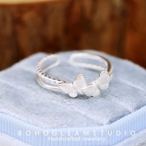 May include: A silver ring with a butterfly design. The ring features two butterflies and a twisted band. The ring is displayed on a light blue velvet surface. The text "BOHOGLEAMSTUDIO Handcrafted Jewellery" is visible.
