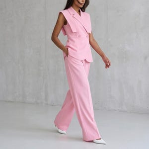 K&ouml;nnte beinhalten: Ein hellrosa &auml;rmelloser Blazer mit steigendem Revers und zweireihiger Knopfleiste, kombiniert mit einer passenden Hose mit weitem Bein. Das Set zeichnet sich durch ein strukturiertes Oberteil mit Schulterdetails und ein schlichtes Design aus.