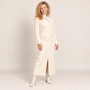 May include: A long, cream-colored coat featuring a high collar, long sleeves, and a front button closure. The coat has a tailored fit with a straight silhouette that extends to the ankles, featuring a central slit at the bottom hem.