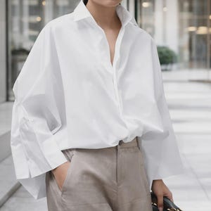 May include: A white, oversized button-down shirt with a classic collar and long sleeves. The shirt is paired with beige trousers. The sleeves are wide and gathered at the cuffs. The shirt is unbuttoned at the top, creating a V-neck.