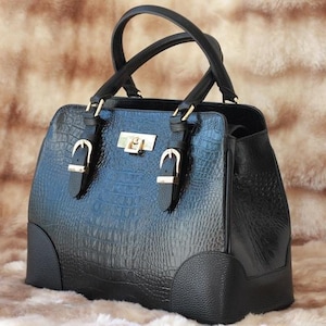 Puede incluir: Bolso de mano negro con un patrón texturizado similar al de un cocodrilo. El bolso tiene dos asas superiores, hebillas doradas y una forma rectangular. La base y los paneles laterales son negros.