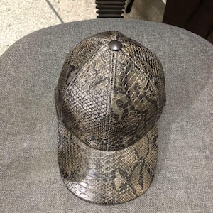 May include: A brown and tan baseball cap with a snakeskin pattern. The cap has a curved brim and a button on top. The hat is sitting on a gray chair.