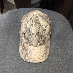 May include: A baseball cap with a snakeskin pattern in shades of beige, gray, and black. The cap has a curved brim and a button on top. The hat is made of a textured material.