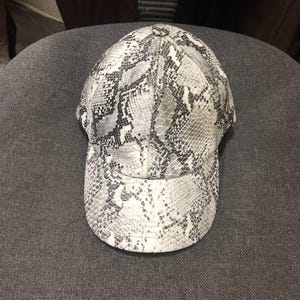May include: A baseball cap with a snakeskin pattern in shades of gray and white. The cap has a curved brim and a button on top. The hat is made of a textured material.