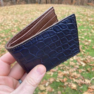 May include: A navy blue wallet with a crocodile skin texture is open, revealing a brown interior with card slots. The wallet is held against a blurred background of fall foliage.
