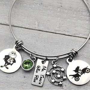 May include: A silver charm bracelet with multiple charms, including a green crystal, a frog wearing a tuxedo, a charm with the text "Mr. Toad's Wild Ride", a charm with a spiral of stars, and a charm with a silhouette of a car with a witch driving.