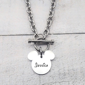 Pode incluir: Um colar de prata com fecho de correr e um pingente em forma de Mickey Mouse com o nome "Jessica" gravado.
