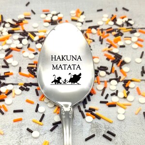 May include: A stainless steel spoon with the words "HAKUNA MATATA" and a silhouette of Simba, Timon, and Pumbaa. The spoon is surrounded by colorful sprinkles in white, orange, and brown.