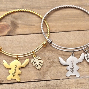 Puede incluir: Dos pulseras de brazalete de oro y plata con dijes. Los dijes son un personaje de dibujos animados con la palabra "Ohana" grabada en &eacute;l y una hoja tropical.