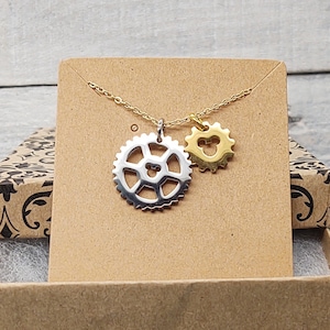 May include: A gold necklace with a silver gear charm and a heart charm. The necklace is displayed on a brown cardboard box with a patterned design.