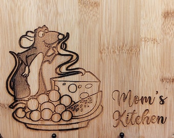 May include: A rectangular bamboo cutting board with a handle. The board features a wood-burned illustration of a cartoon rat, cheese, grapes, and the words "Mom's Kitchen". The board is displayed on a black metal stand.