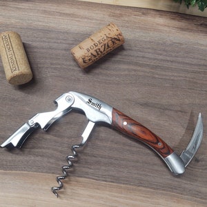 May include: A silver and wood wine opener with a corkscrew, bottle opener, and foil cutter. The opener is engraved with the name "Smith".