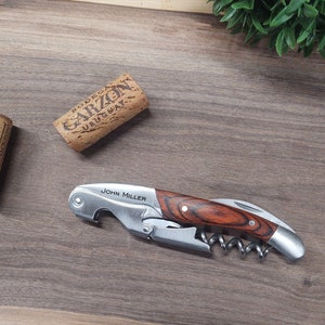 May include: A silver and wood wine opener with a corkscrew, bottle opener, and foil cutter. The opener has a wood handle with a dark brown and light brown wood grain pattern. The handle is engraved with the name "JOHN MILLER".