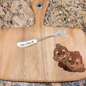May include: A wooden cutting board with a handle, engraved with "Chip 'n Dale" and a small silver spreader. The board features a laser-etched image of Chip and Dale.
