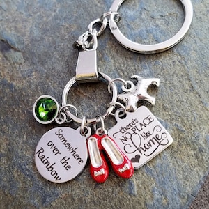 May include: A silver keychain with a green gem, a silver charm of a dog, a silver charm with the text "There's No Place Like Home", and a silver charm with the text "Somewhere over the Rainbow". The keychain also has two red ruby slippers charms.