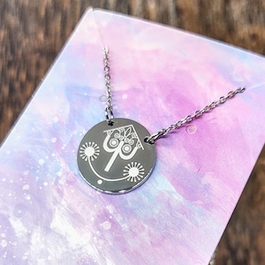 May include: A silver necklace with a round pendant featuring a stylized image of a house with a heart-shaped window and a whimsical design. The pendant is surrounded by small, stylized flowers.