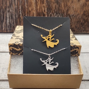May include: Two necklaces with a gold and silver pendant of the character Zero from the movie The Nightmare Before Christmas. The necklaces are displayed on a black card with a white background.