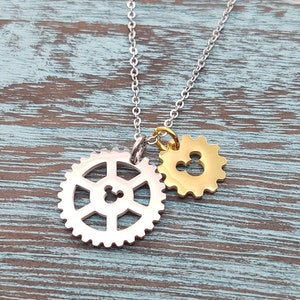 May include: A silver chain necklace with two charms. One charm is a silver gear with a Mickey Mouse silhouette cut out. The other charm is a gold gear with a Mickey Mouse silhouette cut out.