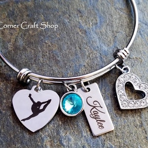 May include: A silver bangle bracelet with four charms: a heart with rhinestones, a heart with a gymnast silhouette, a turquoise blue crystal, and a rectangular tag with the name "Kaylee" engraved on it.