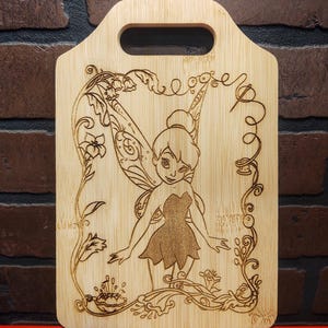 May include: A wooden cutting board with a laser-engraved design of Tinkerbell, the fairy from Disney's Peter Pan. The design features Tinkerbell with her wings spread, surrounded by flowers and vines. The board has a handle on the top.