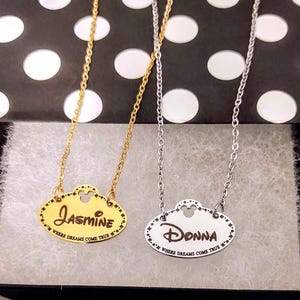 May include: Two oval pendant necklaces on thin metal chains. One pendant is gold-toned with the name Jasmine engraved, and the other is silver-toned with the name Donna engraved. Both feature a small mouse head cutout and the text WHERE DREAMS COME TRUE.
