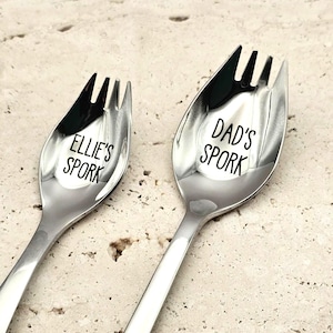 May include: Two polished stainless steel sporks with a shiny finish. The smaller spork is engraved with the text "ELLIE'S SPORK" and the larger spork is engraved with the text "DAD'S SPORK". Both utensils feature a spoon bowl with fork tines.