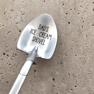 May include: A silver-colored metal spoon shaped like a small shovel with a pointed tip. The bowl of the spoon features the black engraved text DAD'S ICE CREAM SHOVEL. The handle extends downwards from the center of the shovel-shaped bowl.