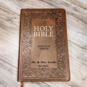 May include: A brown leather-bound Holy Bible with ornate embossed detailing. The cover features the words "HOLY BIBLE" and "KING JAMES VERSION." Personalized with "Mr. & Mrs. Smith Est. 2022."
