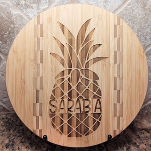 May include: Round bamboo cutting board with a carved pineapple design and the name "SARABIA" engraved below. The board has a checkered pattern around the edge.