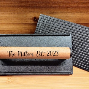 May include: A black cast iron grill press with a wooden handle. The handle is engraved with the text "The Millers Est. 2023".