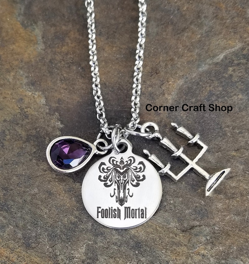 May include: A silver necklace with a charm that says "Foolish Mortal" and features a haunted mansion icon. The necklace also has a purple teardrop-shaped crystal and a silver candelabra charm.