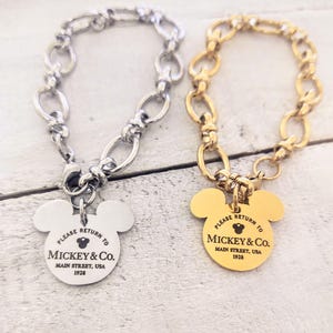 May include: Two Mickey Mouse charm bracelets, one silver and one gold. Both charms feature the words "Please Return To Mickey & Co. Main Street, USA 1928".