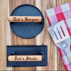 May include: Two cast iron grill presses with wooden handles. The top press has the text "Dad's Burgers" on the handle. The bottom press has the text "Dad's Steaks" on the handle.