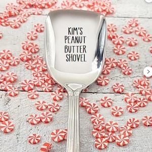 May include: A shiny, silver peanut butter shovel with the text "KIM'S PEANUT BUTTER SHOVEL" printed on the scoop. The shovel is surrounded by red and white peppermint candies on a light-colored wooden surface.