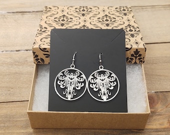 May include: A pair of silver earrings with a detailed design of a spooky mansion. The earrings are displayed on a black card in a brown box with a patterned design.