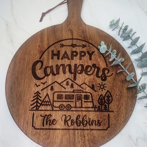 May include: A round wooden cutting board with a brown wood grain. It has a handle and is engraved with the text "Happy Campers" and a camper van, mountains, trees, and a sun. The text "The Robbins" is also engraved on the board.