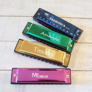 May include: Four harmonicas in different colors, blue, green, gold, and pink, with personalized names on each: Amanda, Timothy, Michelle. The harmonicas have 10 holes and are labeled with numbers 1 through 10.