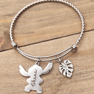Puede incluir: Un brazalete de plata con una banda de metal trenzado. El brazalete tiene dos dijes: un personaje Stitch de plata con la palabra "Ohana" grabada en &eacute;l y un dije de hoja de monstera de plata.
