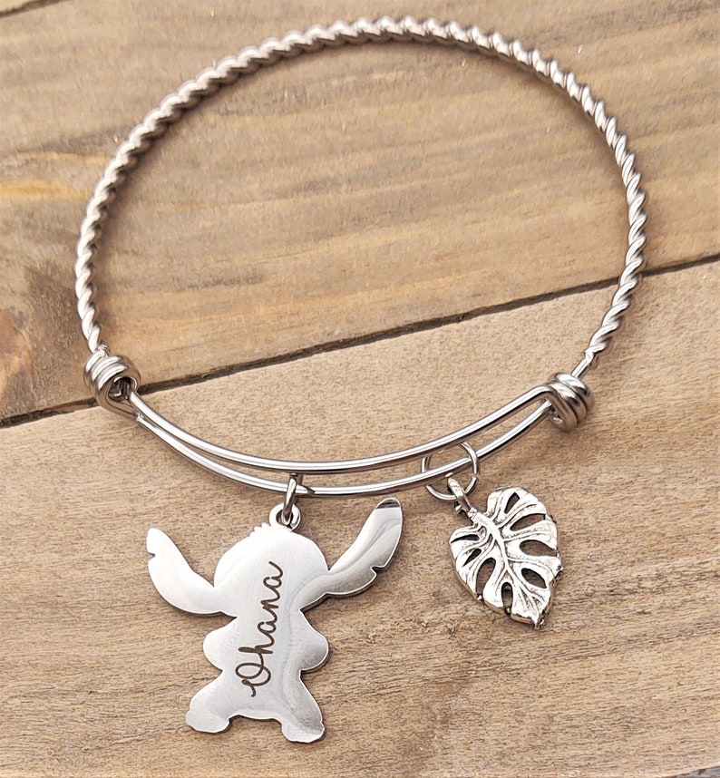 Puede incluir: Un brazalete de plata con una banda de metal trenzado. El brazalete tiene dos dijes: un personaje Stitch de plata con la palabra "Ohana" grabada en &eacute;l y un dije de hoja de monstera de plata.