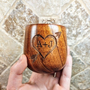 May include: A wooden cup with a heart and arrow design engraved on the side. The heart has the initials "A+J" inside.