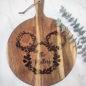 May include: A round wooden cutting board with a handle. The board is engraved with two wreaths and the text "The Millers".