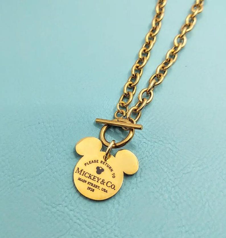 Pode incluir: Colar de corrente dourada com um pingente em forma de cabe&ccedil;a do Mickey Mouse. O pingente tem o texto "Please return to Mickey & Co. Main Street, USA 1928".