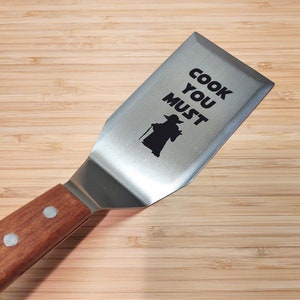 May include: A brown wooden handled spatula with a stainless steel blade. The blade has a black silhouette of Yoda from Star Wars and the text "Cook You Must".